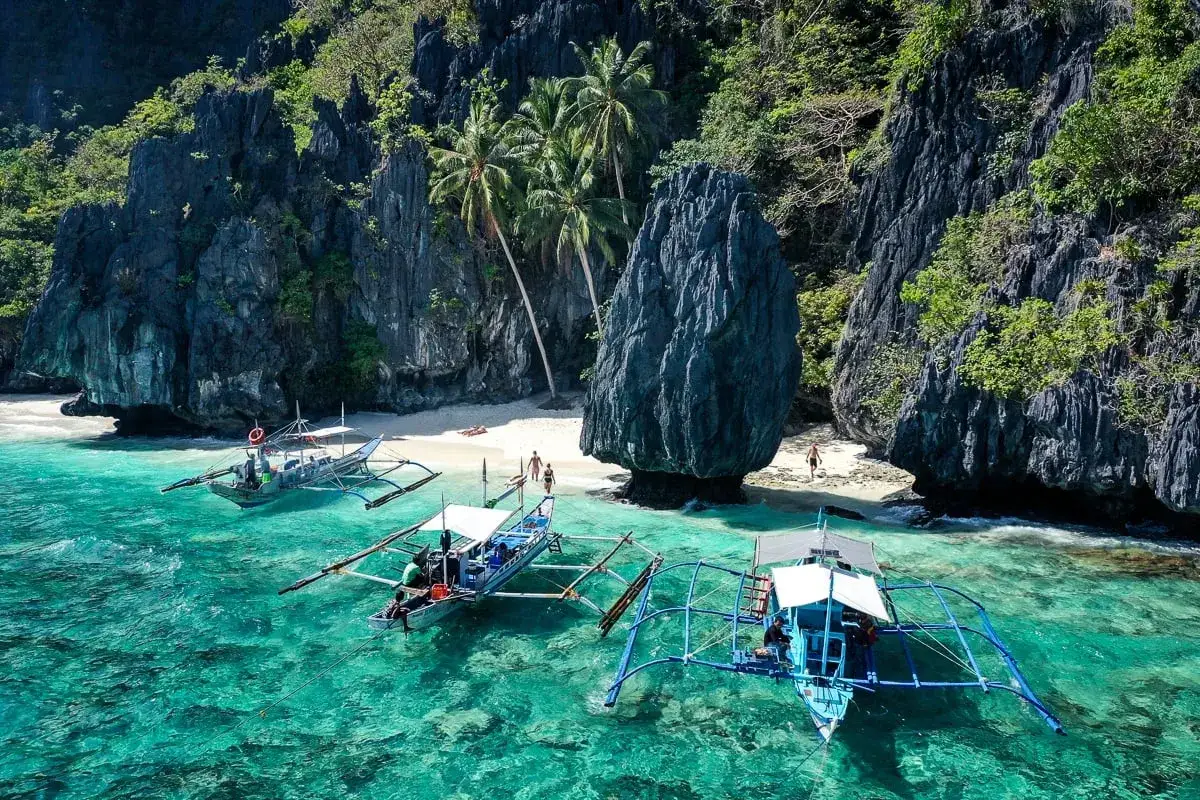 Barcos en aguas turquesas, frente a una playa de arena blanca y acantilados verdes. Un lugar perfecto para saber qué hacer en el nido.