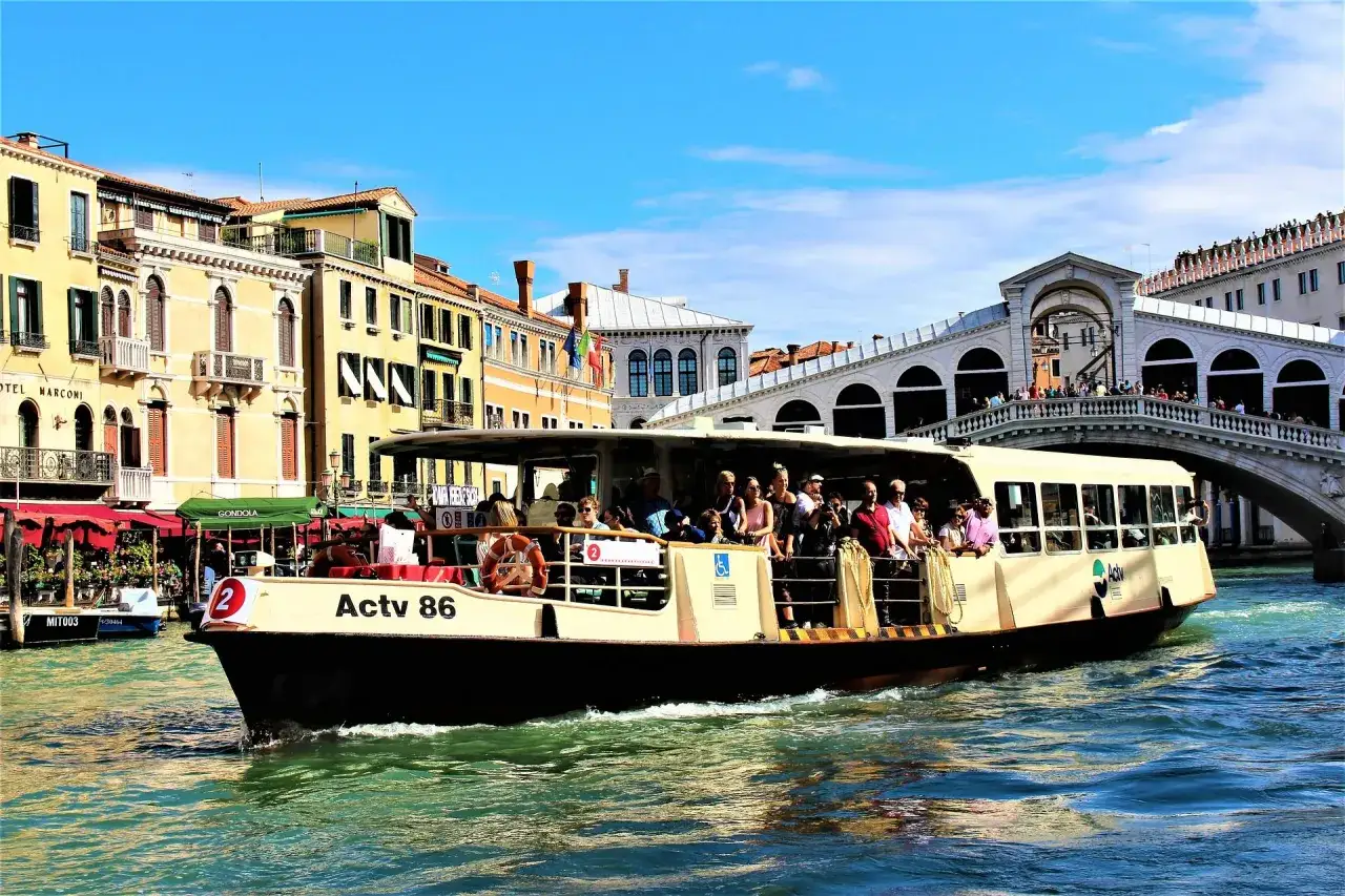 Un vaporetto lleno de turistas navega por el Gran Canal de Venecia, pasando bajo el ic&oacute;nico Puente de Rialto. Una forma ideal de como moverse por Venecia.