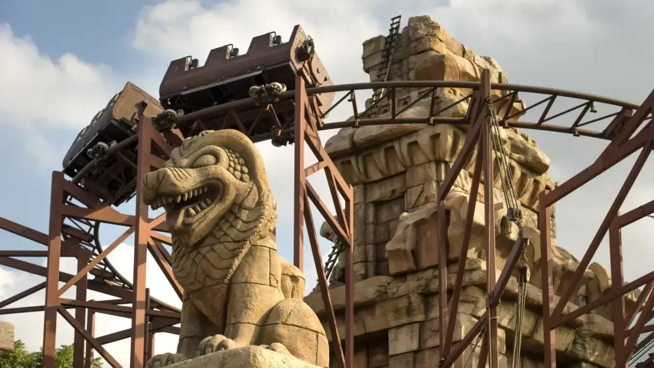 El tren de la montaña rusa disneyland paris se desliza junto a una estatua de león de piedra y ruinas antiguas.