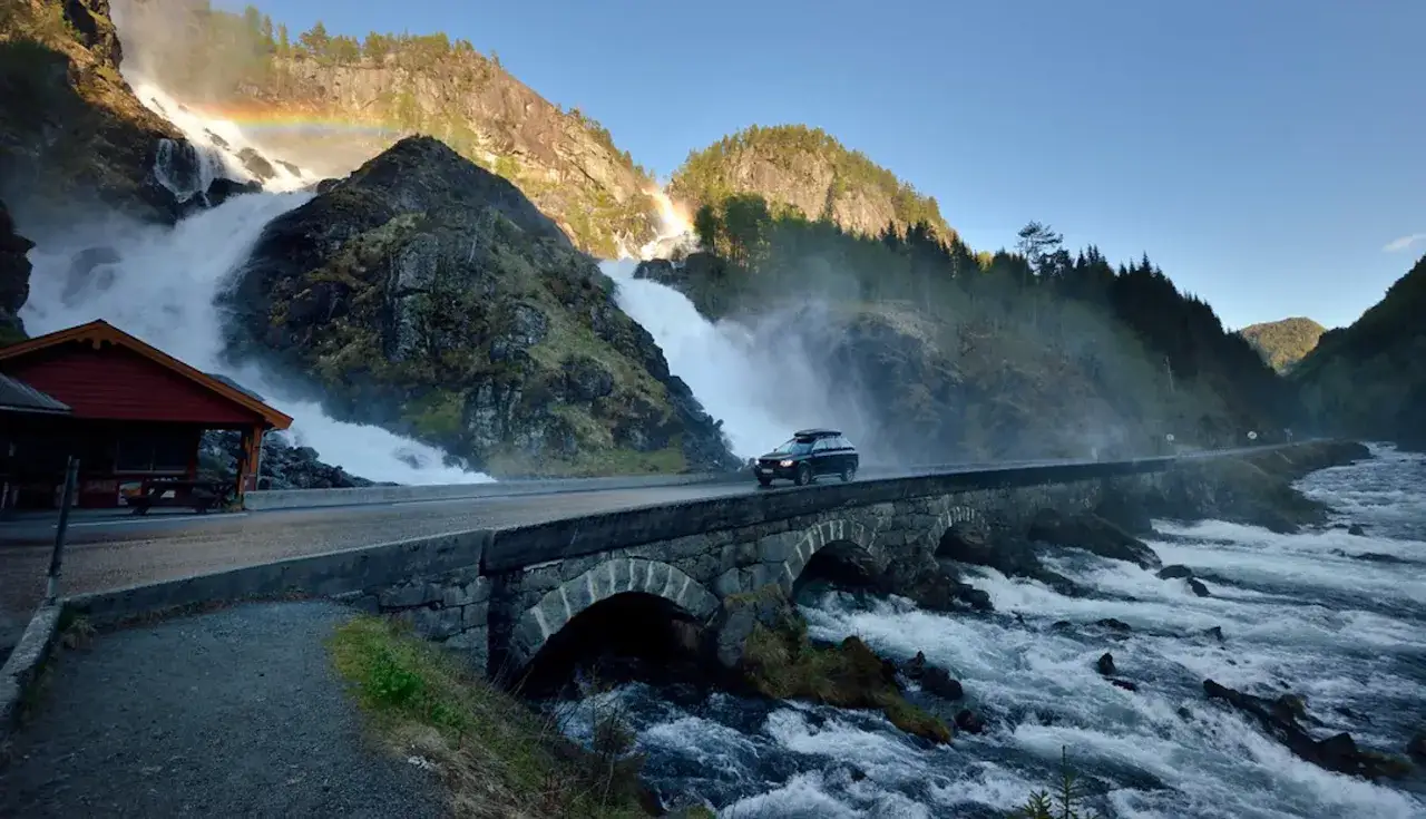 Un coche cruza un puente de piedra sobre un río caudaloso, con una cascada y un arcoíris al fondo. Una aventura de noruega en coche.