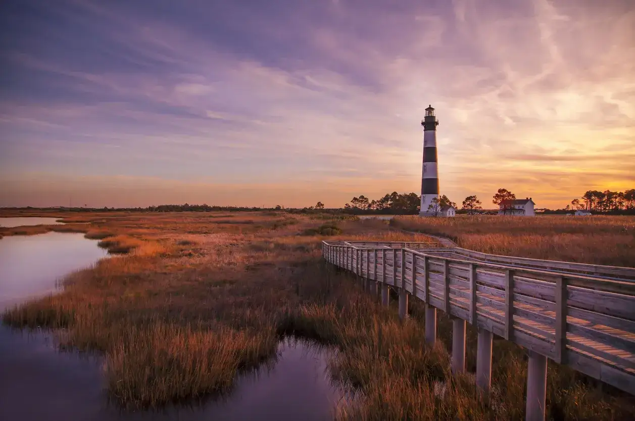 Atardecer en este hermoso outer banks lugar. Un faro ic&oacute;nico se alza sobre marismas doradas, con un camino de madera que invita a explorar.