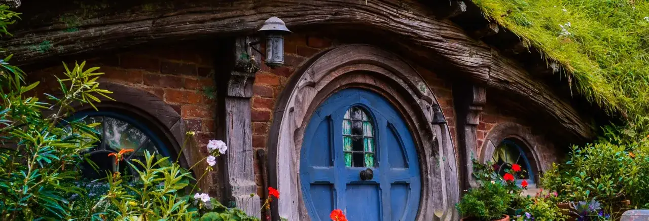 Puerta azul de una casa hobbit en Hobbiton, Nueva Zelanda, rodeada de flores y vegetaci&oacute;n exuberante.