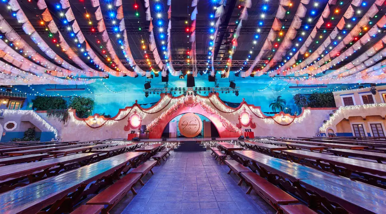 Mesas largas y bancos de madera en la cantina PortAventura, decorada con luces de colores y un escenario festivo.