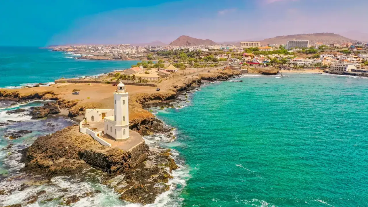 Faro blanco en Cabo Verde, con aguas turquesas y una ciudad costera. El turismo en Cabo Verde puede ser peligroso si no se toman precauciones.