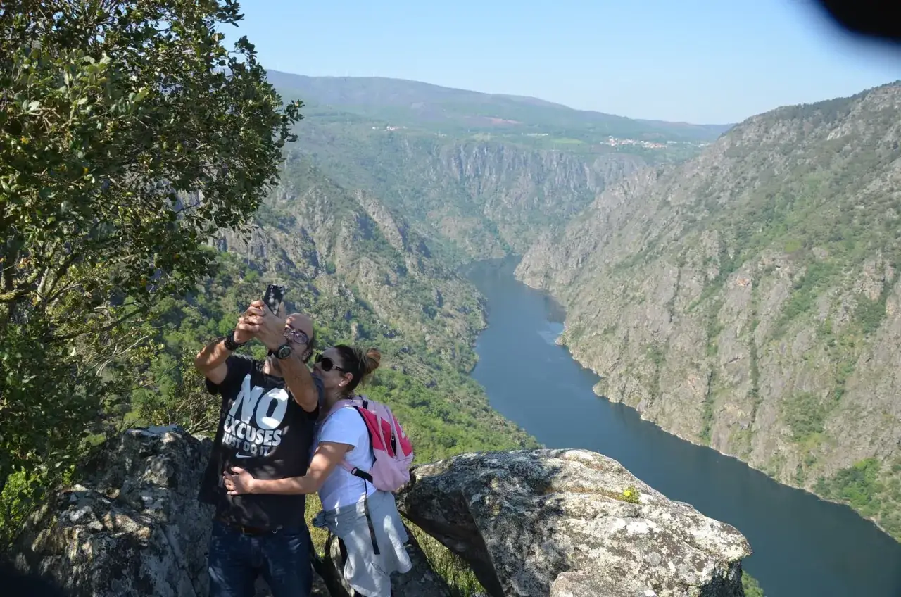 Pareja disfrutando de impresionantes rutas por Galicia, capturando un selfie con un ca&ntilde;&oacute;n y r&iacute;o al fondo.