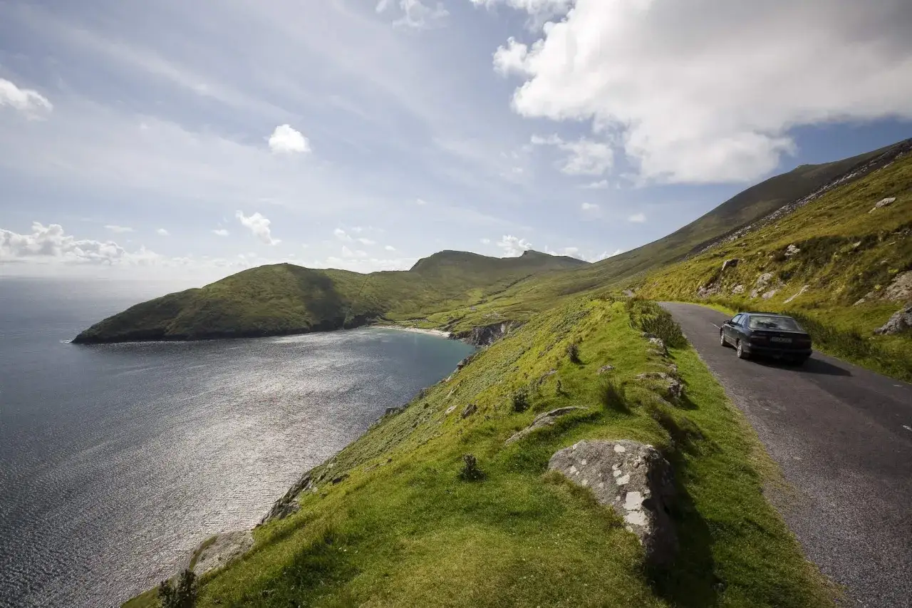 Coche negro en una carretera costera, disfrutando de una ruta por Irlanda. Paisaje verde y mar azul bajo un cielo nublado.