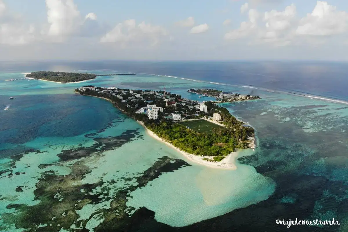 Isla con edificios, campo de fútbol y palmeras, rodeada de aguas turquesas y azules. Un sueño para viajar a Maldivas.