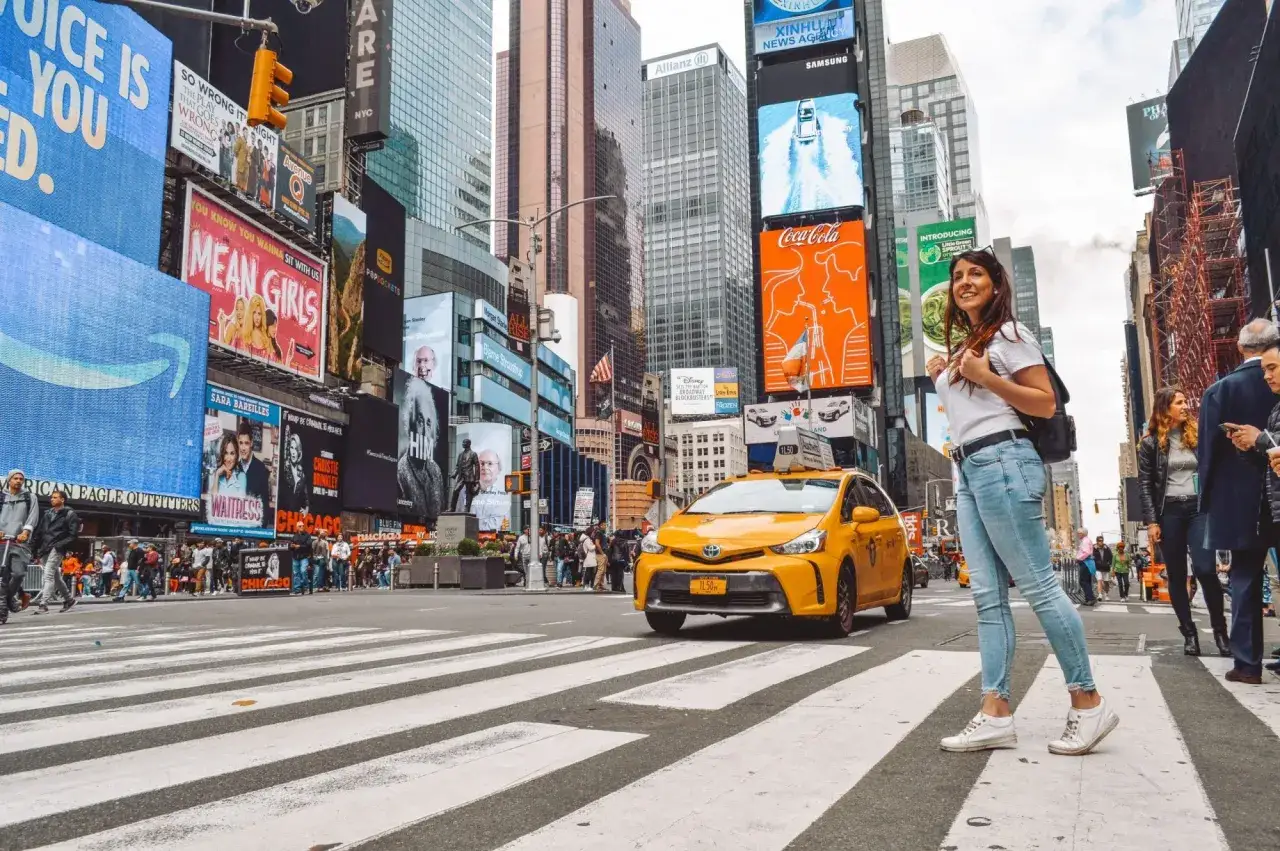 Una mujer sonríe mientras cruza la calle en Times Square, Nueva York, con un taxi amarillo y anuncios brillantes. ¡Una visita a Nueva York inolvidable!