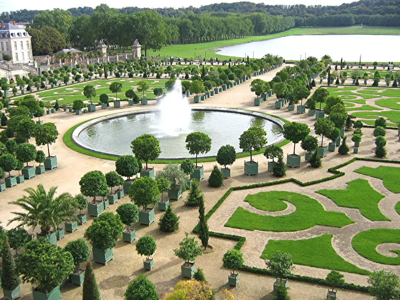 Jardines del palacio de Versalles con fuente central y estanques, árboles en macetas y césped geométrico.
