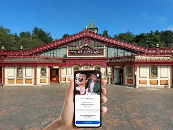 Mano sujetando un teléfono que muestra la opción "Cola virtual" para conocer a Mickey Mouse en Disneyland Paris, frente al edificio "Meet Mickey Mouse".