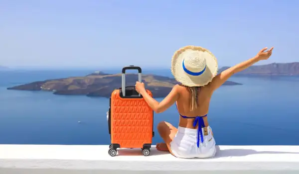 Mujer con sombrero y maleta naranja, lista para decidir dónde viajar en agosto, con vistas al mar y a una isla.