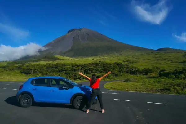 Mujer con los brazos abiertos junto a un coche azul, con el imponente volcán de la isla del Pico de fondo.
