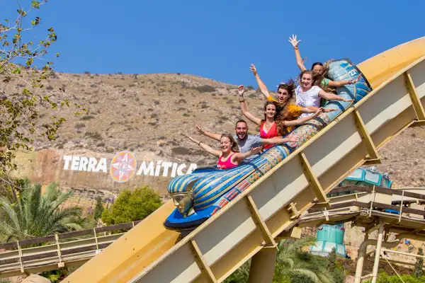 Grupo de amigos ríe y grita en una atracción del parque temático Terra Mítica, con el logo del parque al fondo.