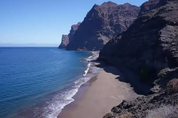 Costa salvaje con acantilados oscuros y una playa de arena dorada bañada por el mar azul. Un rincón secreto entre las mejores playas de Gran Canaria.