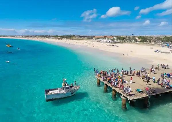 Turistas en un muelle de madera en Cabo Verde, con aguas turquesas y una playa de arena. El turismo en Cabo Verde puede ser peligroso si no se toman precauciones.