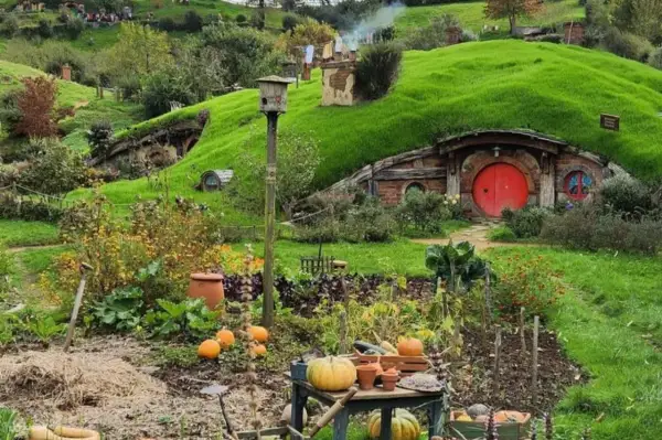 Un jardín exuberante en Hobbiton, Nueva Zelanda, con calabazas y flores, y una casa hobbit con puerta roja.