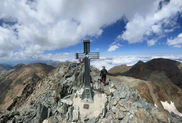 Cima de montaña con cruz metálica. Una persona camina por la ruta fácil, rodeada de picos y nubes.